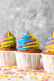 Photo of Delicious cupcakes with colorful cream and sprinkles on white table against blurred grey background, closeup