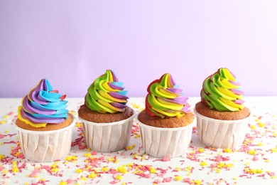 Photo of Delicious cupcakes with colorful cream and sprinkles on white wooden table against violet background