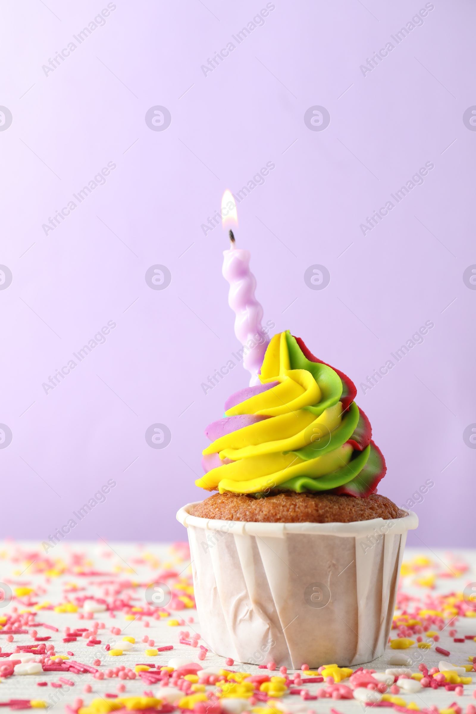 Photo of One delicious cupcake with colorful cream, burning candle and sprinkles on white table against violet background, closeup