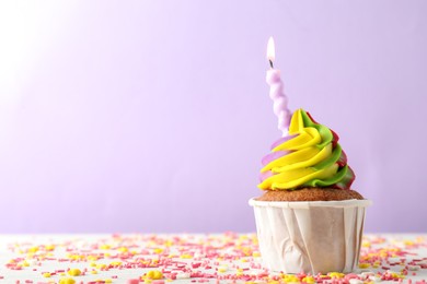 Photo of Delicious cupcake with colorful cream, burning candle and sprinkles on white table against violet background, closeup. Space for text