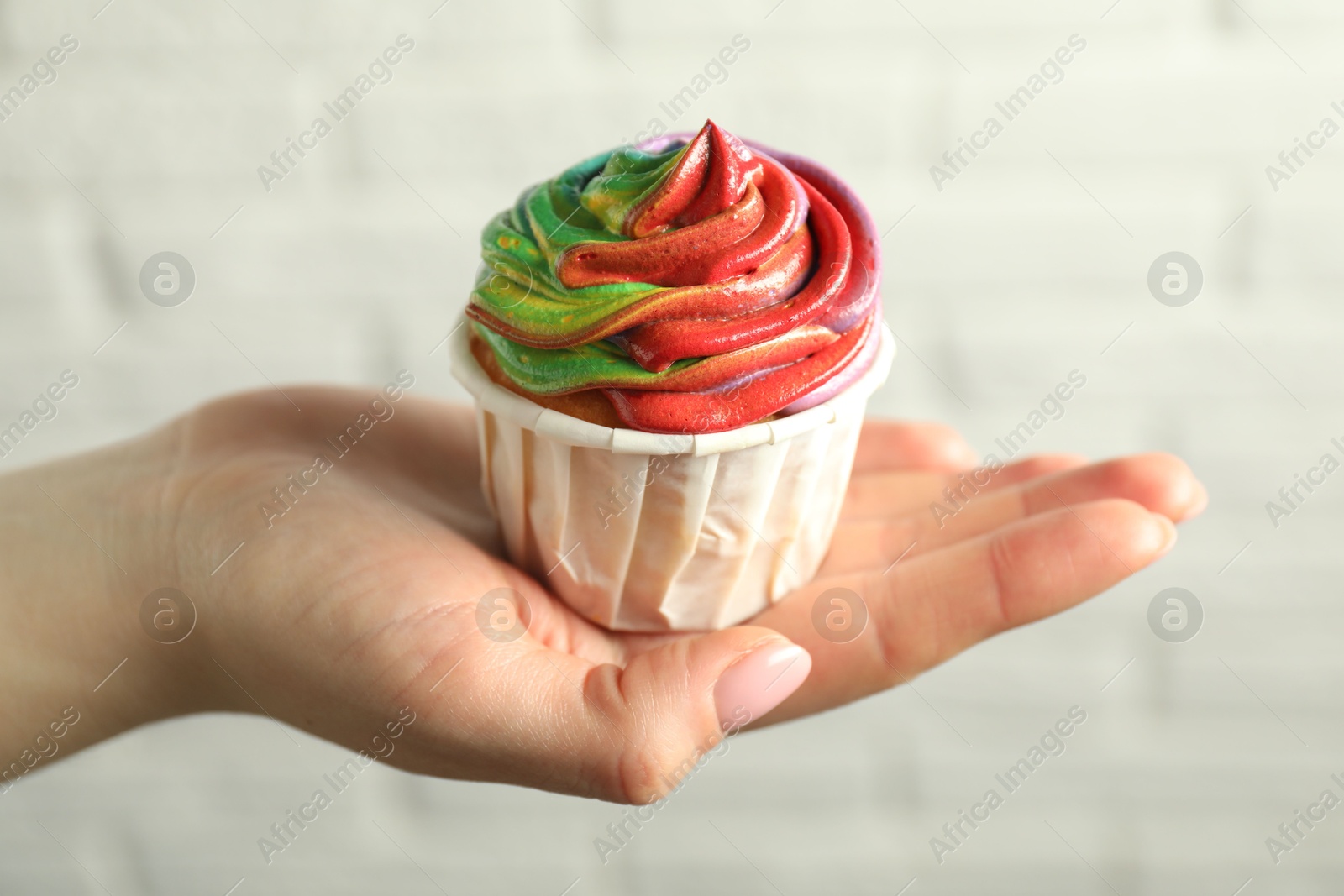 Photo of Woman holding delicious cupcake with colorful cream against white brick wall, closeup