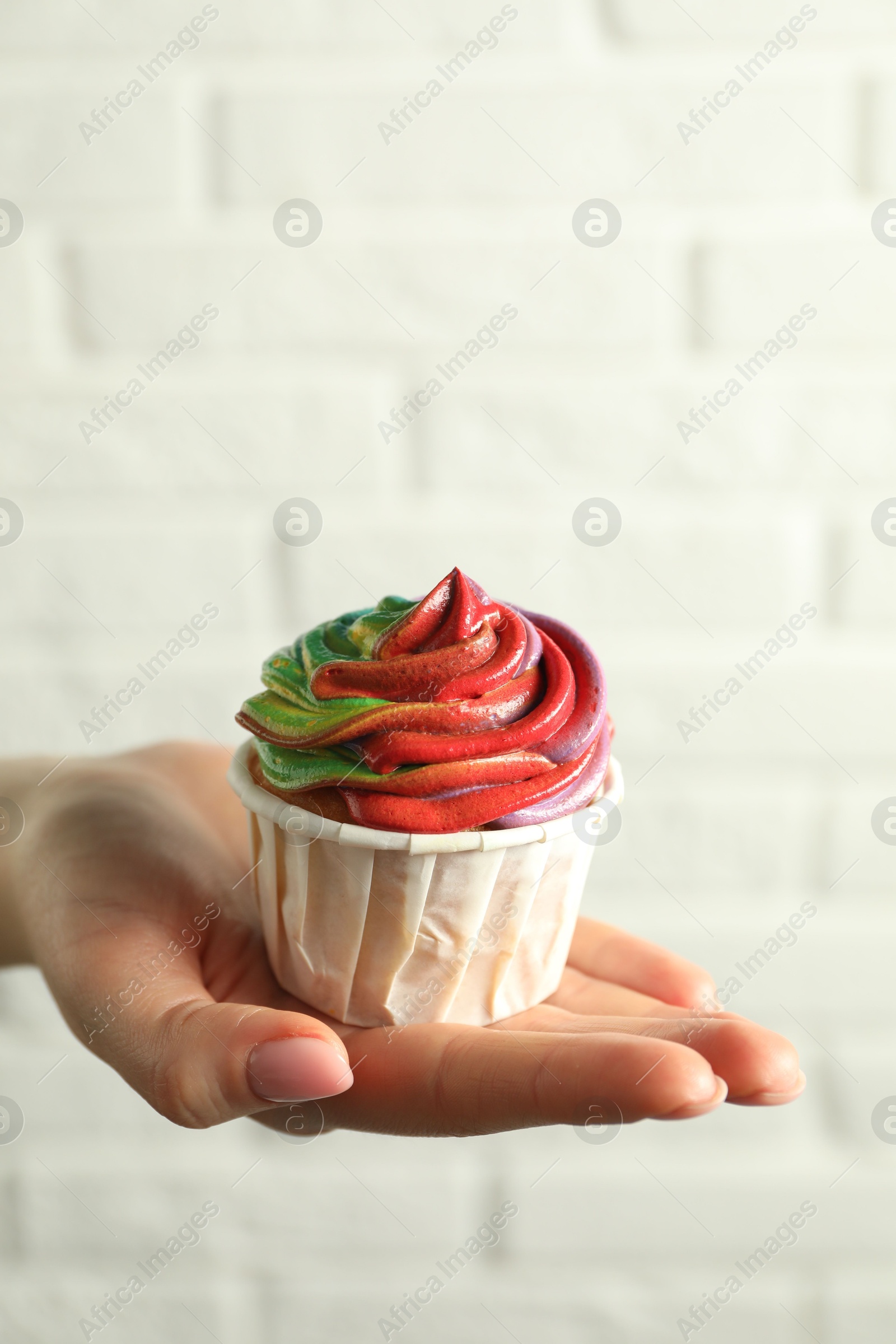 Photo of Woman holding delicious cupcake with colorful cream against white brick wall, closeup. Space for text