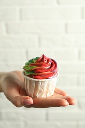 Photo of Woman holding delicious cupcake with colorful cream against white brick wall, closeup. Space for text