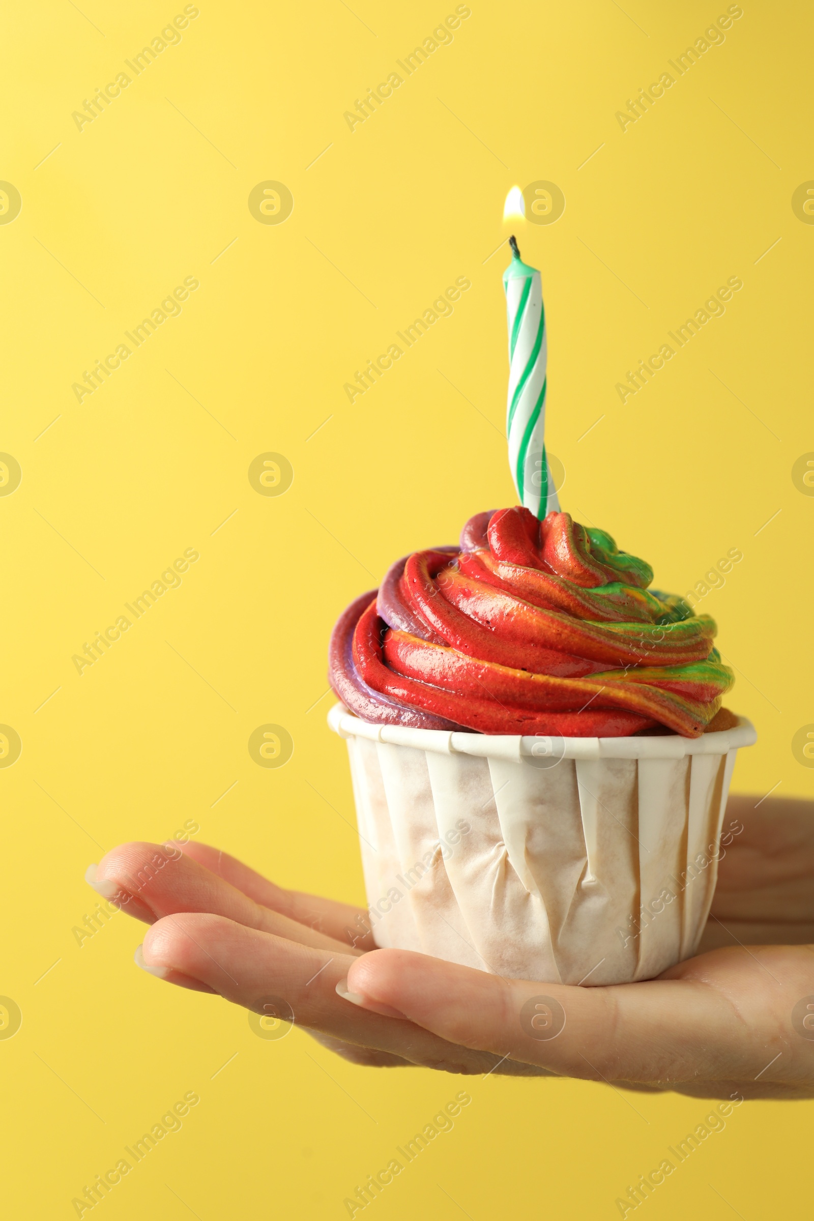 Photo of Woman holding delicious cupcake with colorful cream and burning candle on yellow background, closeup