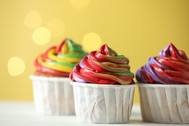 Photo of Delicious cupcakes with colorful cream on white table against golden background, closeup. Bokeh effect