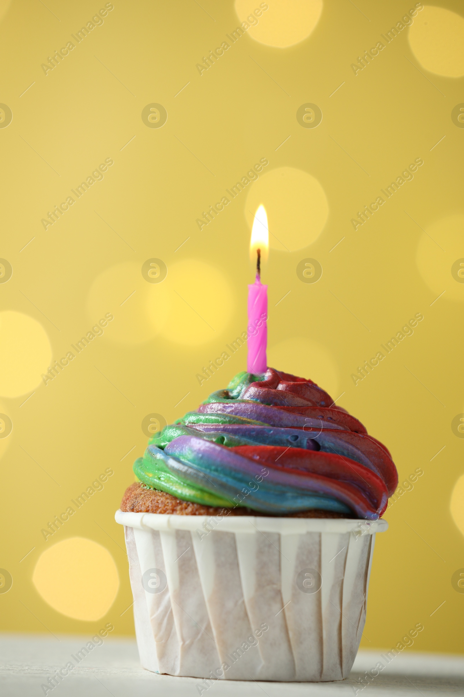 Photo of Delicious cupcake with colorful cream and burning candle on white table against golden background, closeup. Bokeh effect