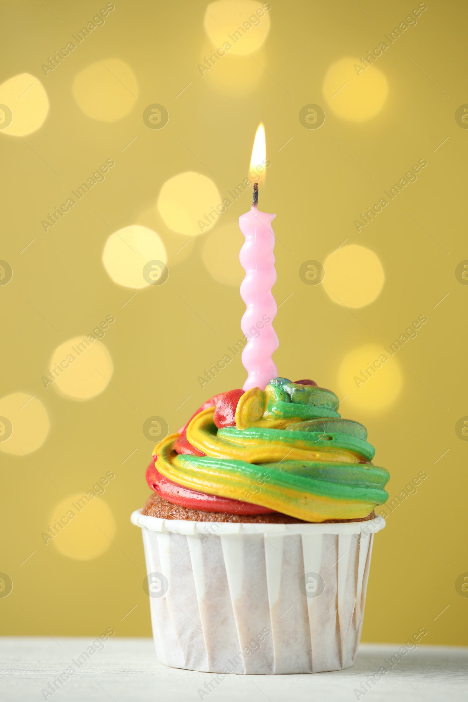 Photo of Delicious cupcake with colorful cream and burning candle on white table against golden background, closeup. Bokeh effect