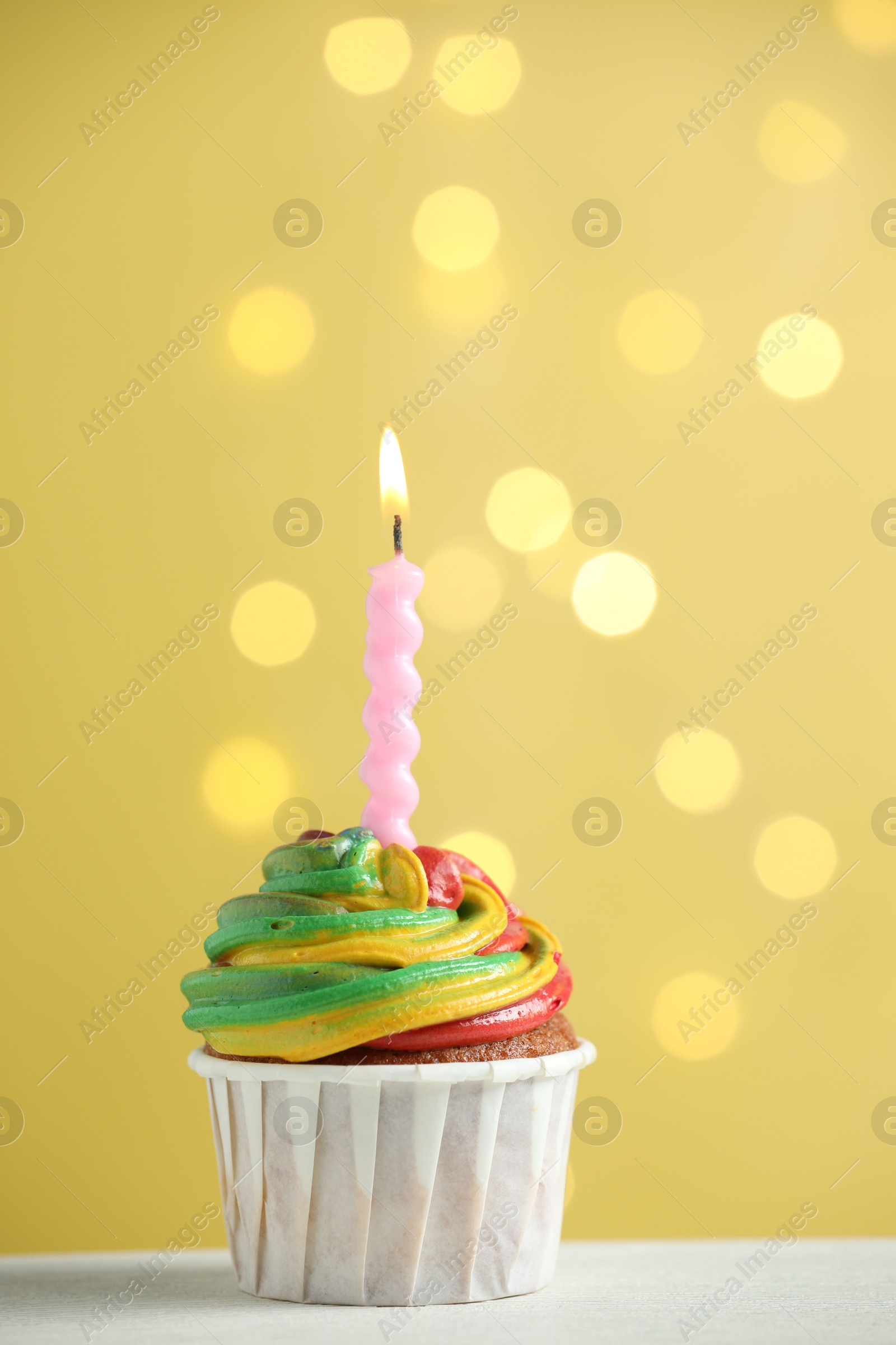 Photo of Delicious cupcake with colorful cream and burning candle on white table against golden background, closeup. Bokeh effect