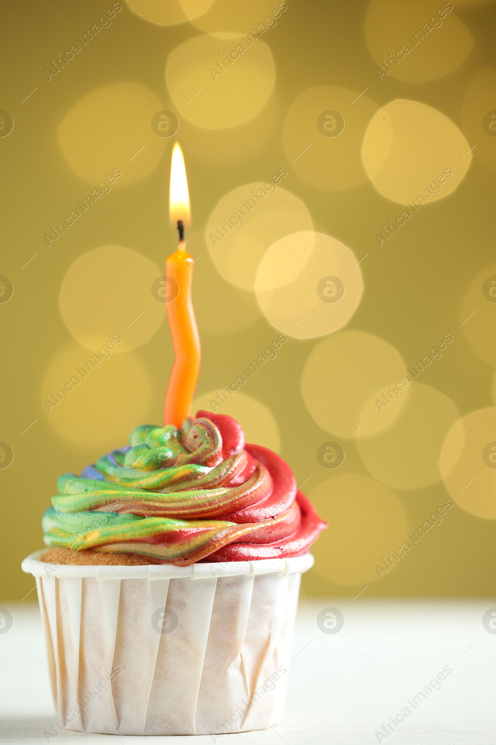 Photo of Delicious cupcake with colorful cream and burning candle on white table against golden background, closeup. Bokeh effect