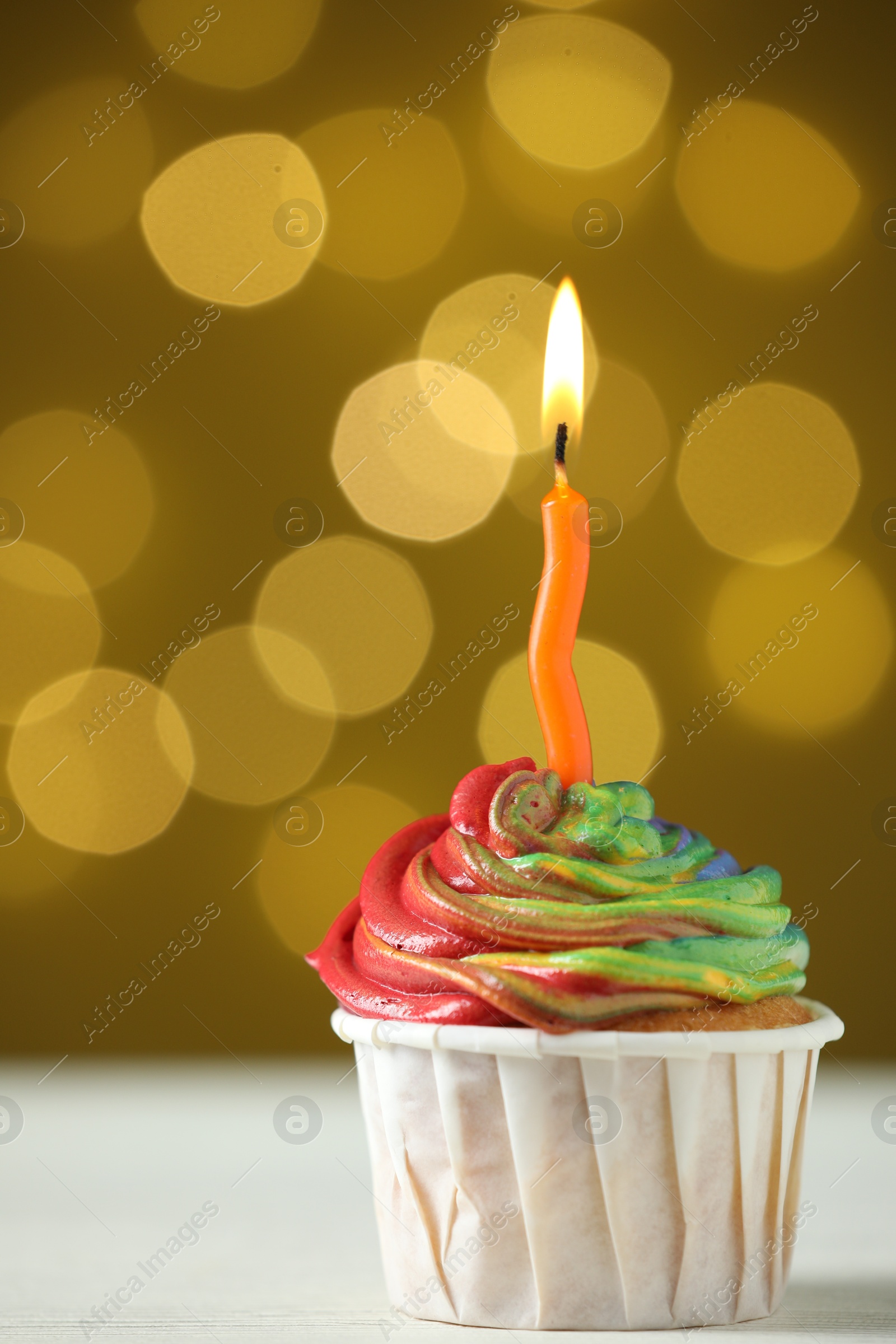 Photo of Delicious cupcake with colorful cream and burning candle on white table against golden background, closeup. Bokeh effect