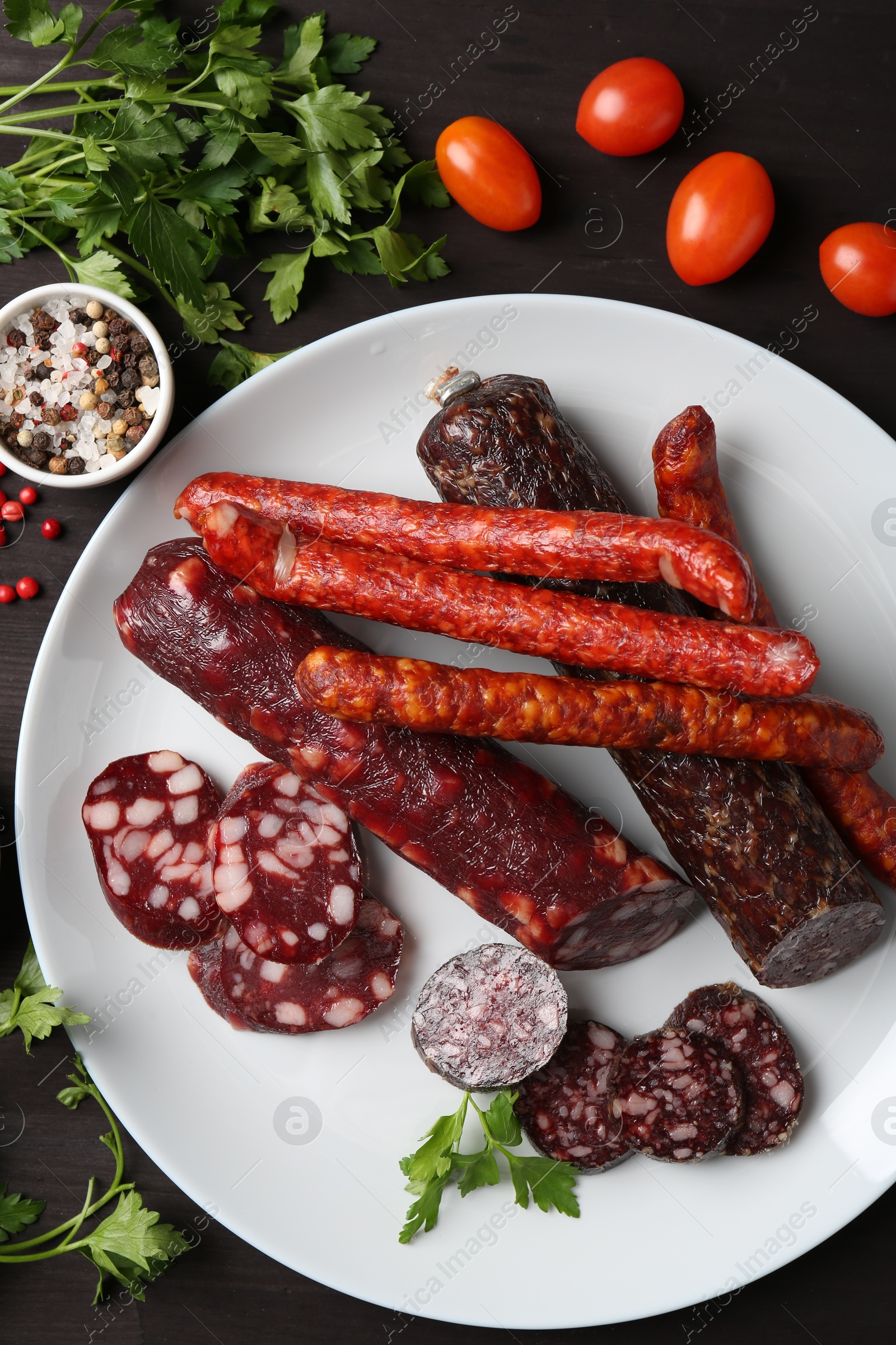 Photo of Different smoked sausages, cherry tomatoes and spices on dark wooden table, flat lay