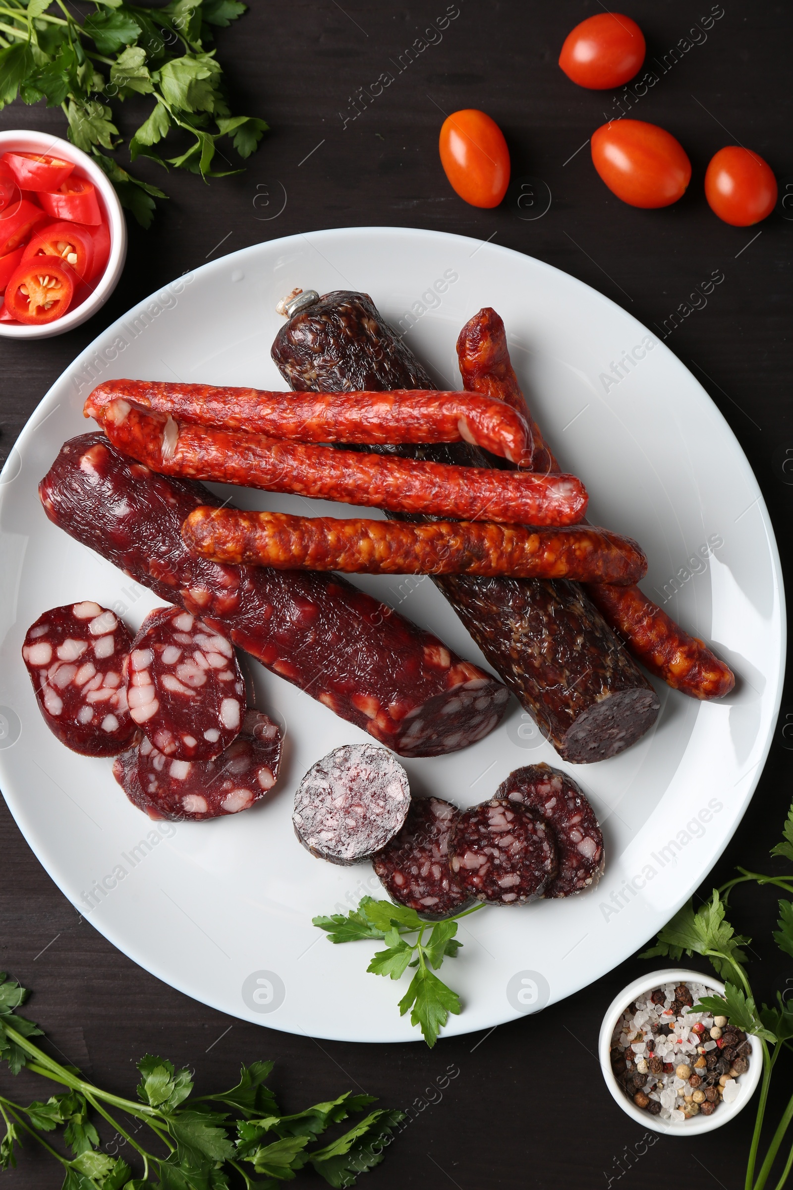 Photo of Different smoked sausages, cherry tomatoes and spices on dark wooden table, flat lay