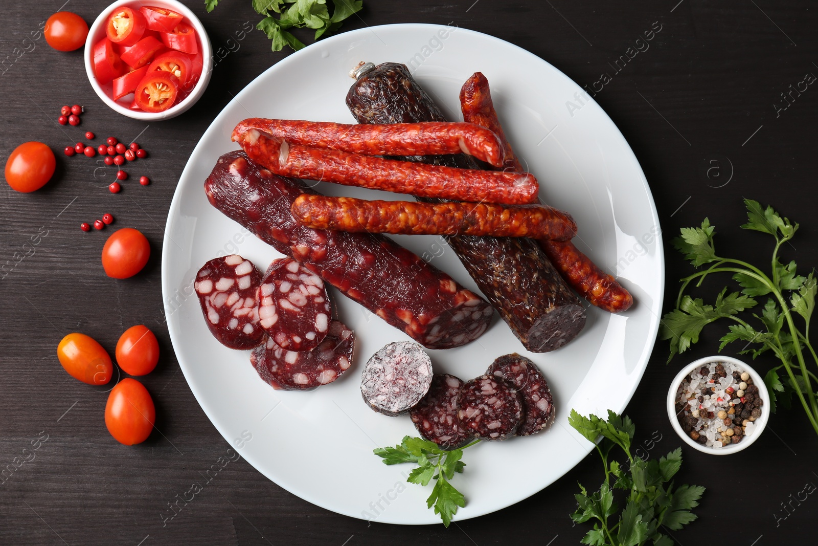 Photo of Different smoked sausages, cherry tomatoes and spices on dark wooden table, flat lay