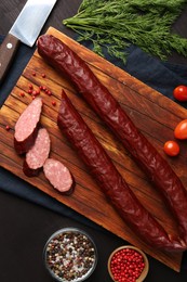 Photo of Delicious thin smoked sausage, cherry tomatoes, spices and knife on dark wooden table, flat lay