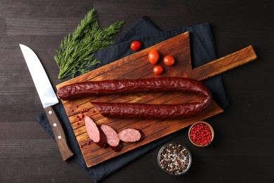 Photo of Delicious thin smoked sausage, cherry tomatoes, spices and knife on dark wooden table, flat lay