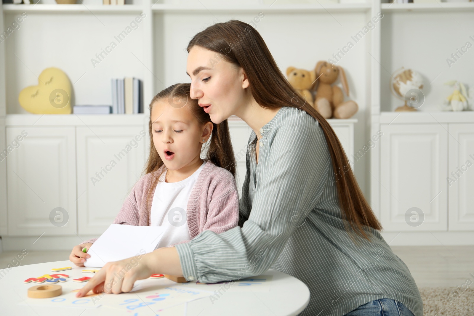 Photo of Speech therapist teaching little girl alphabet at white table indoors