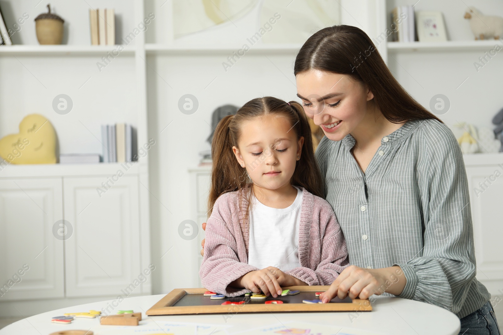 Photo of Speech therapist teaching little girl alphabet with magnetic letters at white table indoors