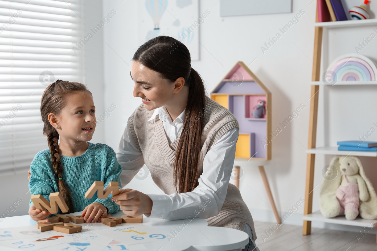 Photo of Speech therapist teaching little girl alphabet with wooden letters at white table indoors