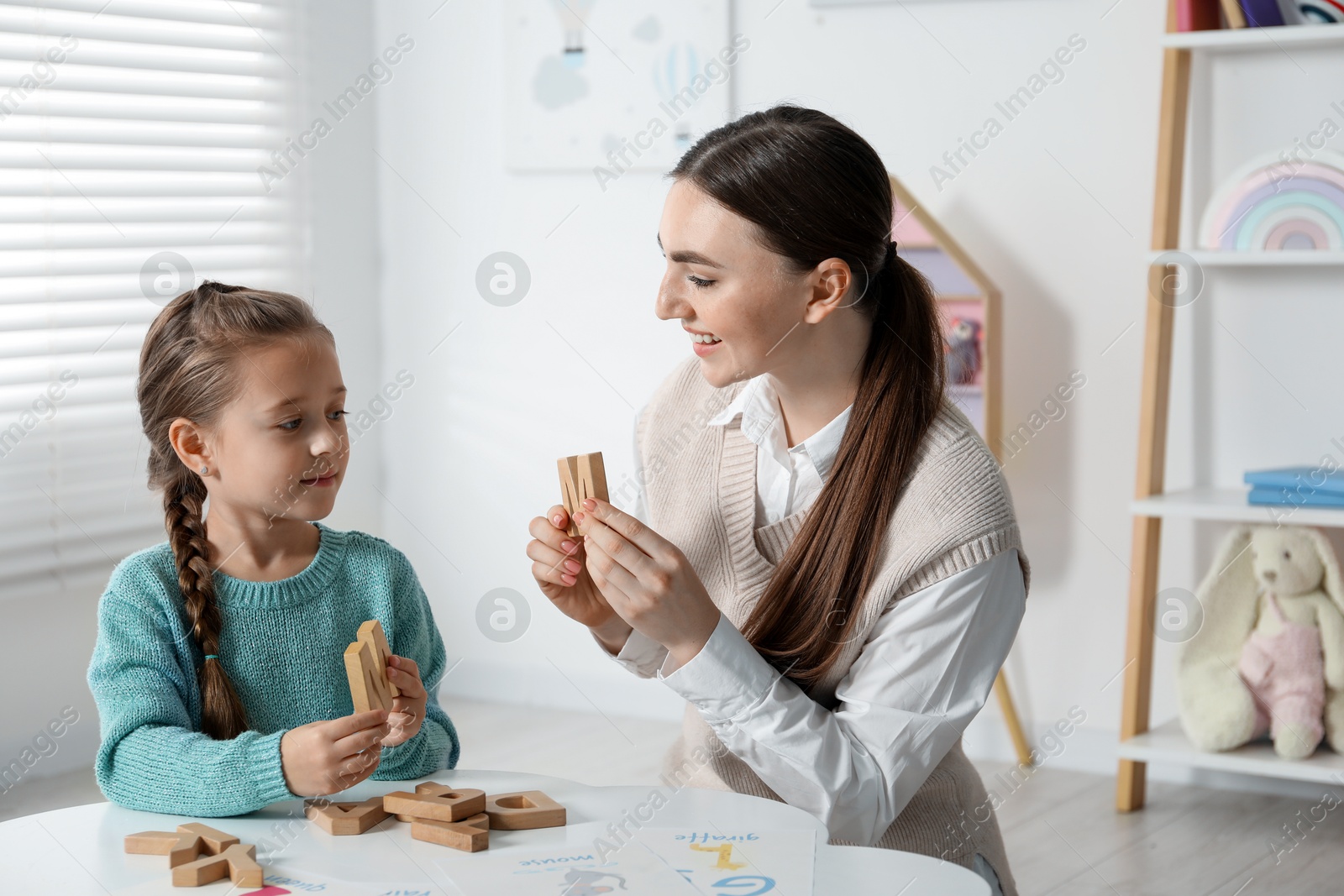 Photo of Speech therapist teaching little girl alphabet with wooden letters at white table indoors