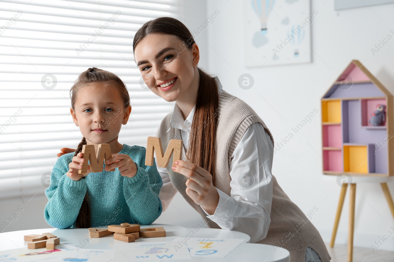 Photo of Speech therapist teaching little girl alphabet with wooden letters at white table indoors