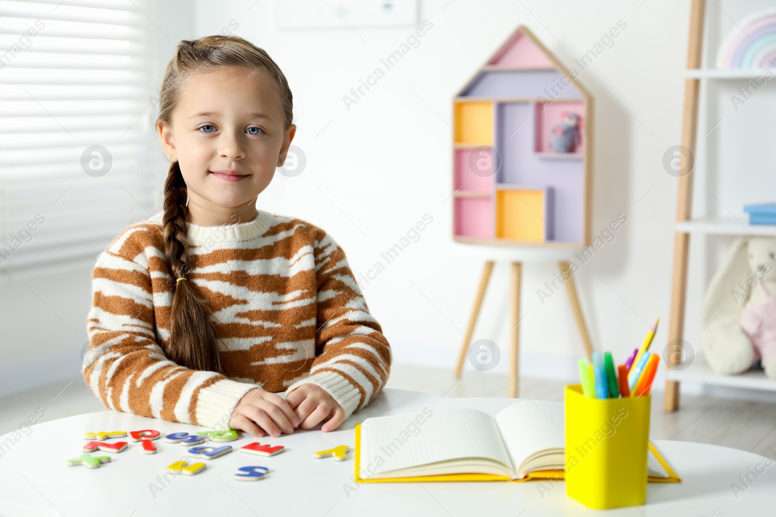 Photo of Little girl learning alphabet with magnetic letters at white table indoors