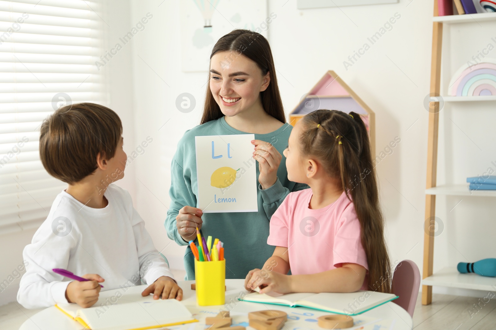 Photo of Speech therapist teaching little kids alphabet at white table indoors