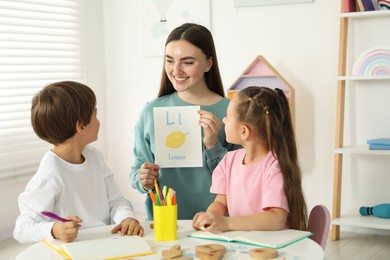 Photo of Speech therapist teaching little kids alphabet at white table indoors