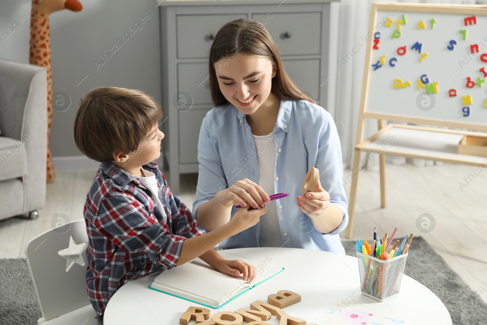 Photo of Speech therapist teaching little boy alphabet with wooden letters at white table indoors