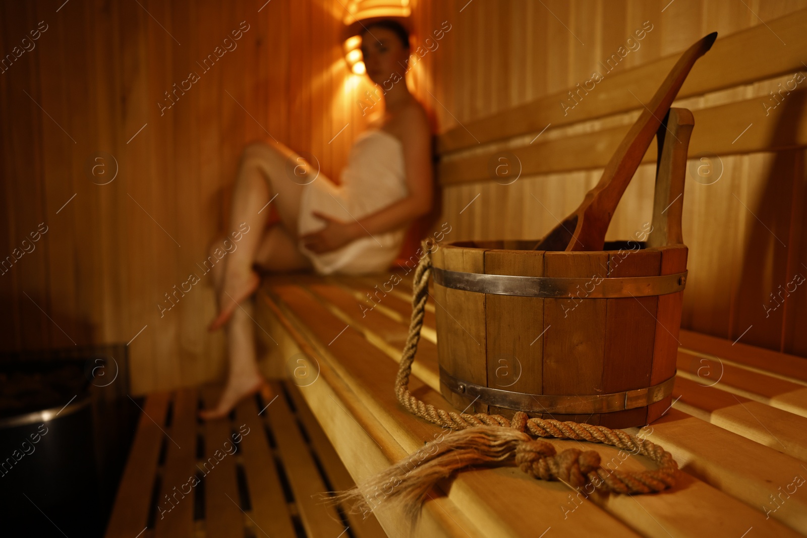 Photo of Bucket with ladle and woman on wooden bench in sauna, selective focus