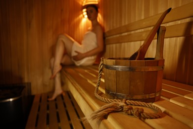 Photo of Bucket with ladle and woman on wooden bench in sauna, selective focus