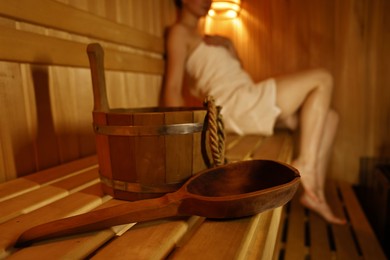 Photo of Bucket with ladle and woman on wooden bench in sauna, selective focus