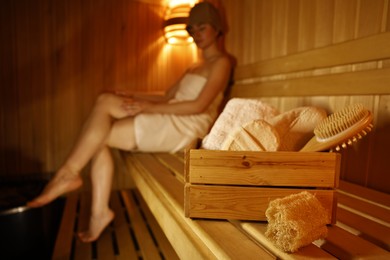 Photo of Crate with bath accessories and woman on wooden bench in sauna, selective focus