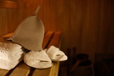 Photo of Felt wool hat, towels and slippers on wooden bench in sauna. Space for text