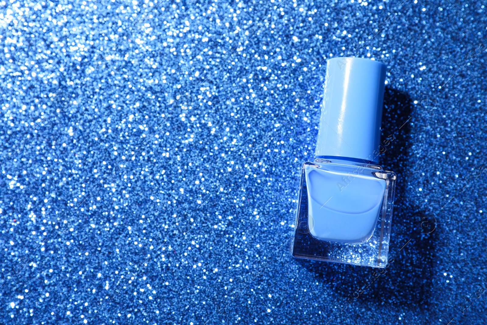 Photo of Nail polish in bottle on blue glitter surface, top view. Space for text