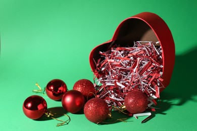 Photo of Red heart shaped box with Christmas balls and shiny filler on green background. Space for text