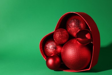 Photo of Red heart shaped box with Christmas balls on green background. Space for text