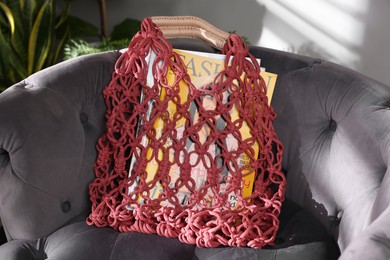 Photo of Handmade macrame shopping bag with magazines on armchair indoors