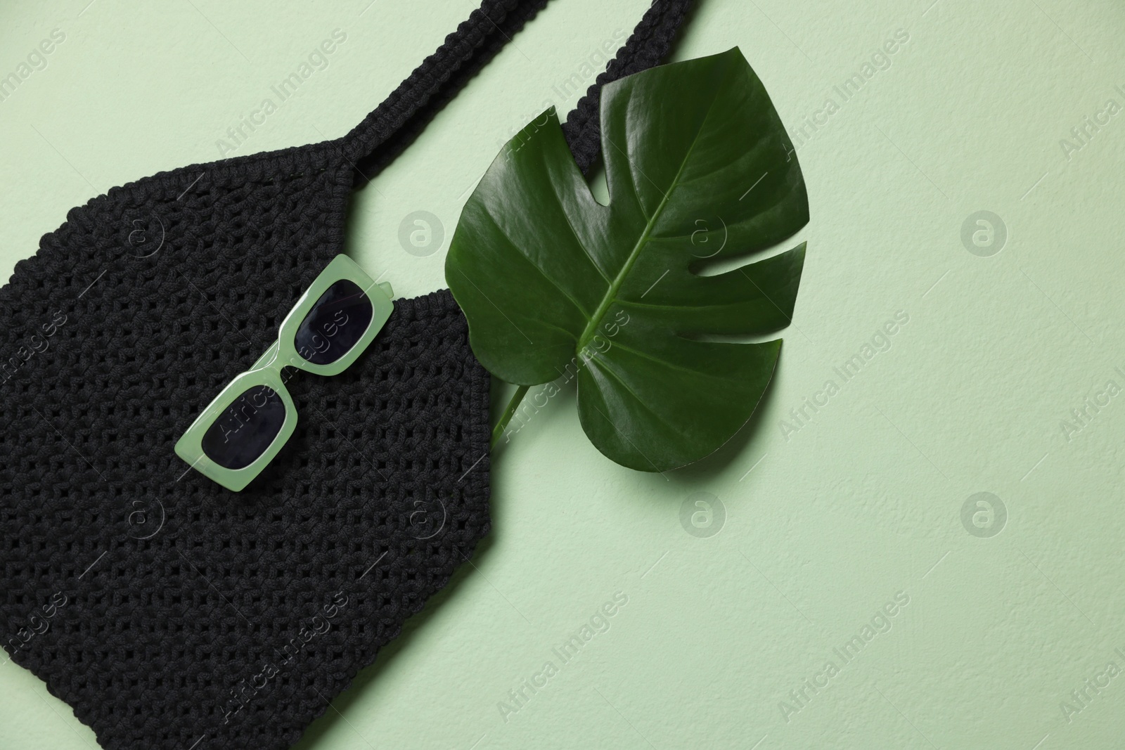 Photo of Handmade macrame shopping bag with leaf and sunglasses on light green background, top view. Space for text