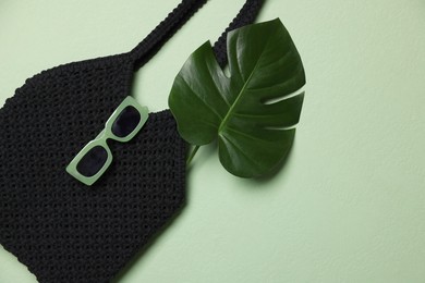 Photo of Handmade macrame shopping bag with leaf and sunglasses on light green background, top view. Space for text