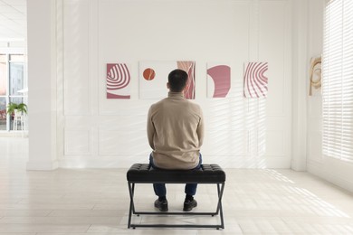 Photo of Man sitting on bench and looking at paintings in art gallery, back view