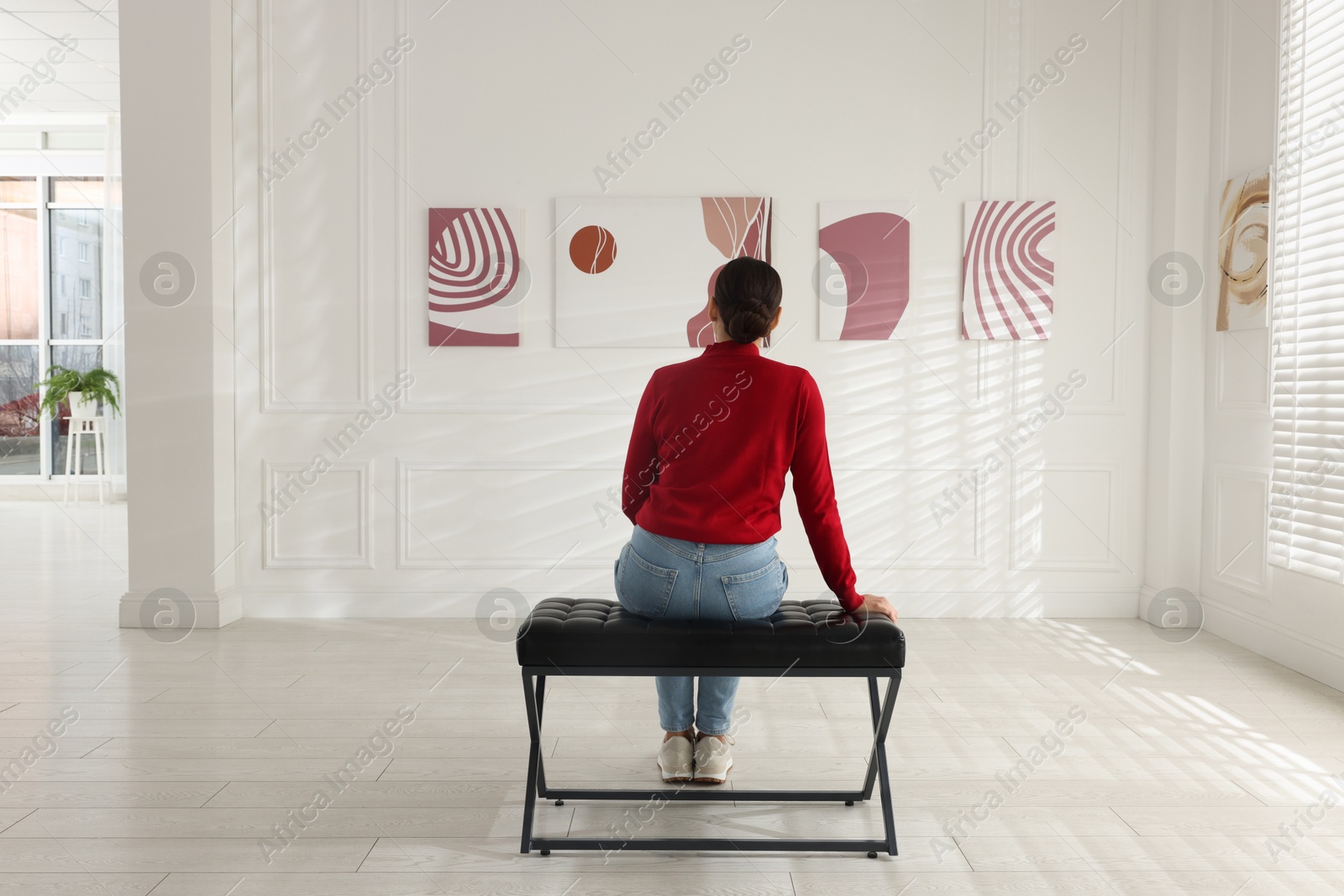 Photo of Woman sitting on bench and looking at paintings in art gallery, back view