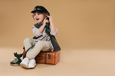 Cute little boy with old telephone and suitcase on beige background, space for text