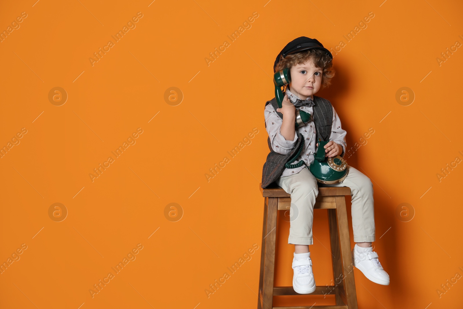 Photo of Cute little boy with old telephone on stool against orange background, space for text