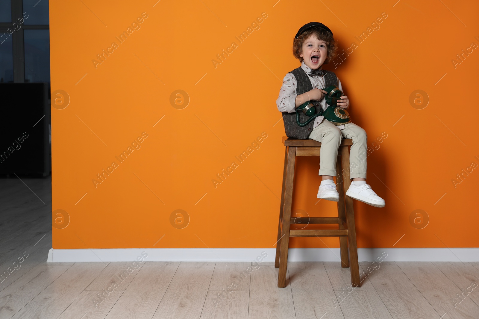 Photo of Cute little boy with old telephone on stool near orange wall, space for text