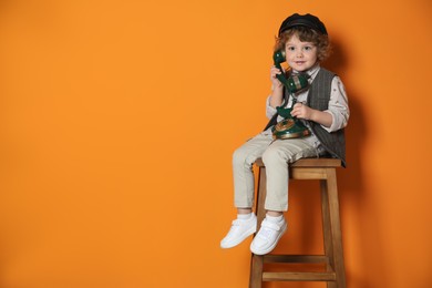 Photo of Cute little boy with old telephone on stool against orange background, space for text