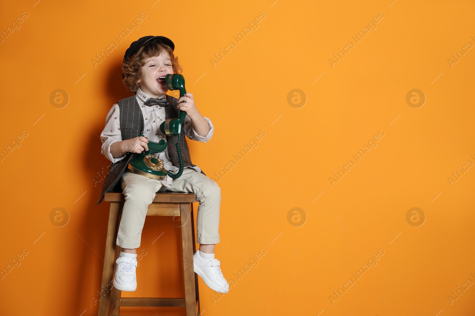 Photo of Cute little boy with old telephone on stool against orange background, space for text