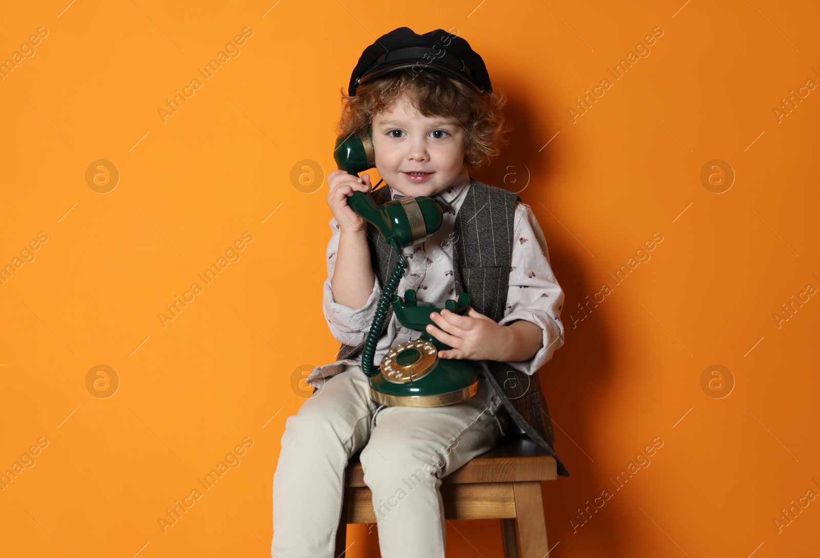 Photo of Cute little boy with old telephone on stool against orange background