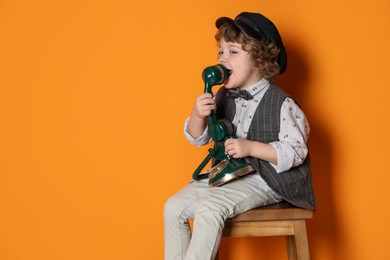 Photo of Cute little boy with old telephone on stool against orange background, space for text