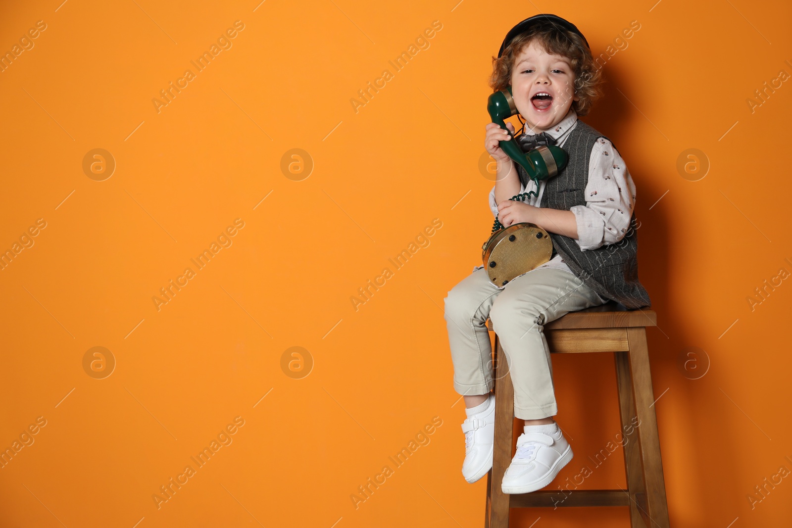 Photo of Cute little boy with old telephone on stool against orange background, space for text