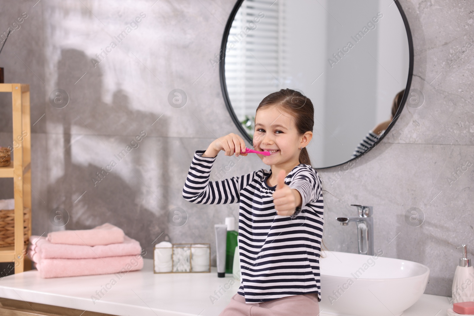 Photo of Cute girl brushing her teeth and showing thumbs up on countertop in bathroom, selective focus